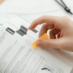 A hand places a sticky note in an open book, symbolizing studying or learning.