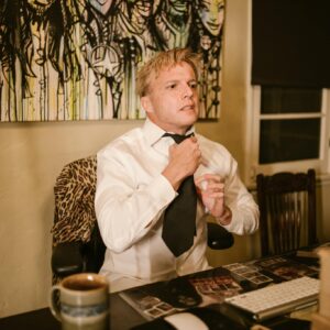 An adult man adjusts his tie in a home office, ready for a virtual meeting, with a cup of coffee on the desk.